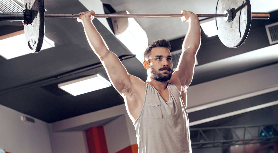 Muscular man performing highly effective strength exercises with the overhead barbell carry and overhead barbell press exercise for bigger muscles