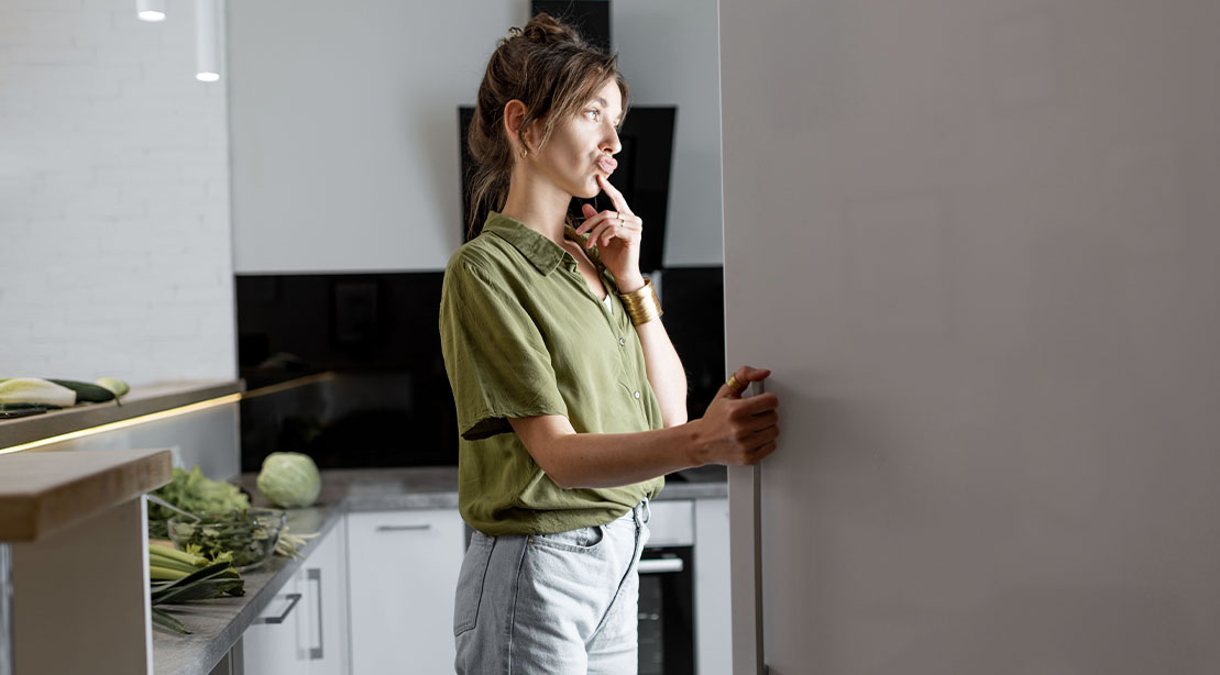 Slim female standing in front of the fridge suffering from food hunger and cravings trying decide what to eat